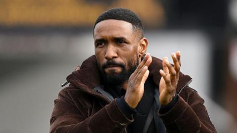 Jermaine Defoe walks across pitch clapping hands, wearing winter unzipped brown jacket