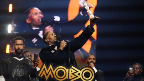Scorcher stands on stage holding a microphone in one hand and an award in the other. He is holding the award in the air. There are four other men on the stage behind him. Two of them are filming him on their mobile phones.