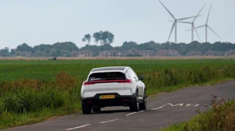 A white electric SUV driving along a country road. There are fields on either side and wind turbines in the distance.