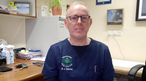 Dr John Diamond, who has thinning grey hair and glasses and who is wearing navy medical scrubs with his name on the chest, sits at a desk with a computer and some papers on it. 