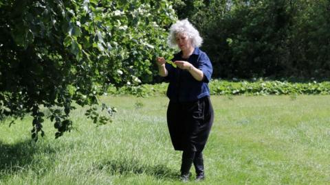 An open meadow with a large tree occupying the left side of the frame and a grassy field stretching across the foreground and background. A woman stands beneath the branches of the tree. She has curly grey hair and is wearing a dark, short-sleeved top, dark trousers, and boots. She is examining leaves from the tree, with both hands raised at chest height.