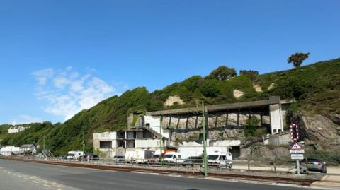 The Summerland site from across the road, you can see the remaining part of a building beneath a cliff, there are some cars parked in front.