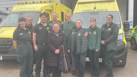 Staff at South East Coast Ambulance Service pose for a photograph with Antony Linward. The group are stood in front of two bright yellow ambulances, with the medical team wearing dark green NHS uniforms.