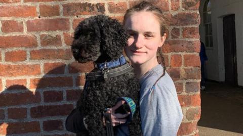 Cerys Lupton-Jones holding a dog. She is a young woman with light brown hair tied into a thin ponytail and wearing a grey shirt. She is standing in front of a brick wall and holding a medium-sized black curly haird dog