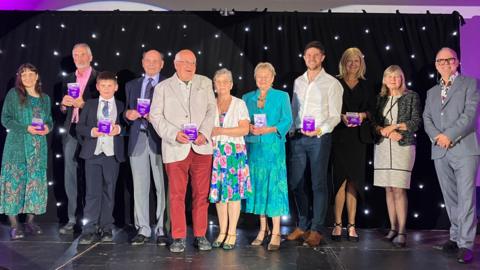 A group of people stand in a line on a stage, and are looking at the camera and smiling. They are holding purple awards in their hands and the background is a black backdrop with white lights shining on it.
