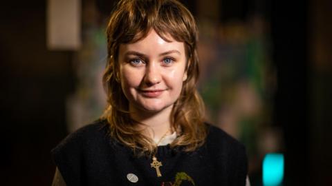 A woman wearing a dark knitted sweater with a cross necklace, standing indoors with soft, colourful, out‑of‑focus lights in the background.