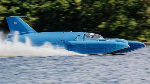 A close-up image showing Bluebird running on Loch Fad. It is a blue craft that looks like a cross between a boat and a plane. It has a tail fin and its pilot can be seen through a clear canopy. Spray is rising up behind it.