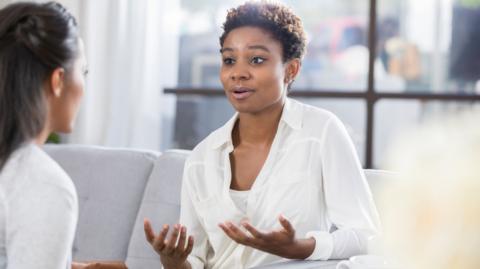 Picture of a women wearing a white shirt speaking to another woman in a white top in an office environment. 
