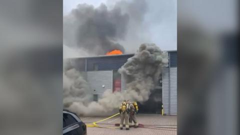 Two firefighters standing in front of an industry unit which has dark smoke coming from the front. There is also an orange blaze coming from the roof.