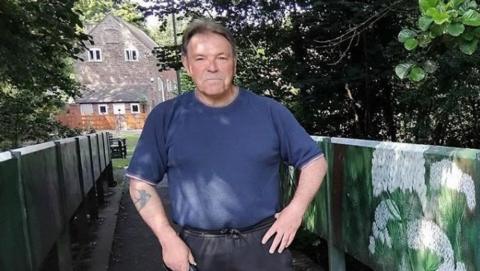Councillor Phil Millward stands at one of the bridges over Hurley Brook. he is wearing a blue T-shirt and it is sunny. There are trees and a house in the background.
