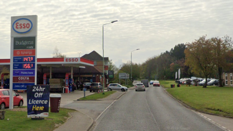 The A143 in Haverhill. An Esso petrol station can be seen on the left hand side of the image. Vehicles can be seen moving ahead in the distance. 