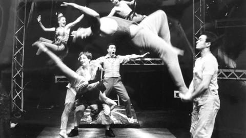 A group of physical theatre performers jumping, doing acrobatics and performing aerial manoeuvres. Rigging can be seen around them. It is an old black and white photograph.