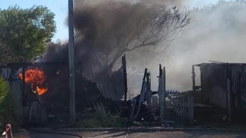 Charred remains of a fence are silhouetted against the grey smoke as flames and black smoke continue to billow out of a substation.
