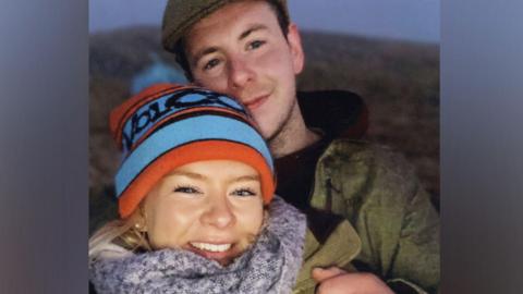 Dr Caroline Muirhead, wearing a blue and red woolly hat and a grey scarf, with Alexander 'Sandy' McKellar standing behind her, wearing a cap and dark jacket