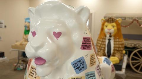 The image shows three lion sculptures. Two in the background are slightly blurred and the one if the foreground is in focus. The one in the front is white and painted with pink heart eyes. It has a number of different coloured squares which each say different messages about Portsmouth. The one in the background is painted in to look like it is wearing a suit. It has a brown jacket on and a black tie. It has a ginger mane and a brown hat on. The third lion is inspired by history of libraries. It is a brown lion, which has green and grey on it.