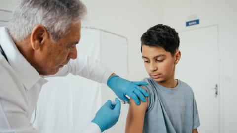 A teenge boy wearing a grey t-shirt receives a vaccine in his arm from a doctor wearing a white coat and blue latex gloves
