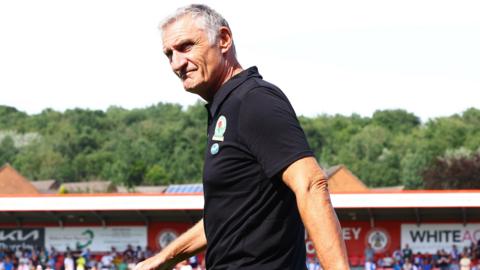 Blackburn Rovers manager Tony Mowbray walks across the pitch with a stand and trees at Accrington Stanley's Wham Stadium in the background.