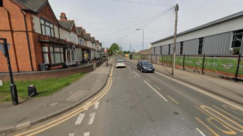 A generic image of Aston Lane, going through the middle of the photo, from the bottom going upwards. Two cars are on the road. Housing is to the left of the street and another building is to the right.