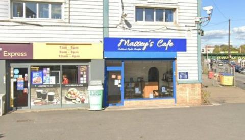 A parade of shops shows Massey's Cafe at the end of the row, which has a blue sign and yellow lettering that promotes the premises. There is a large pavement in front of the cafe. It is a sunny day.