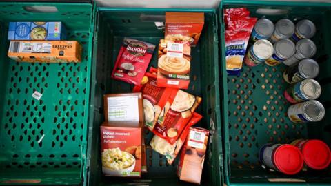 Three large green crates with tins and packets of food in.