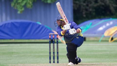 A batsman wearing a blue and yellow kit goes to run after playing a ball to point. 