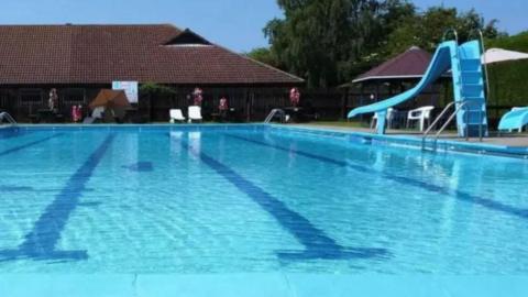 An outdoor swimming pool, with dark blue tiles at the bottom of the pool which mark out the lanes. To the right of the pool there is a blue slide. In the background are chalet-style buildings.