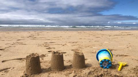 Three small sandcastles and a blue sand bucket and spade on a sandy beach with cloudy sky over the sea