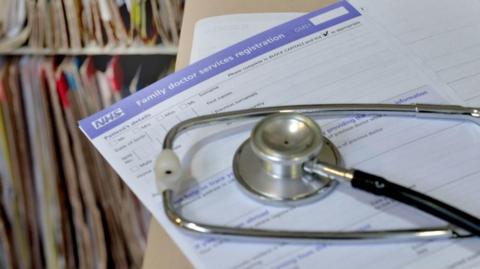 A stethoscope set on an NHS family doctor form. The form is on top of a beige file while in the background is two shelves full of files.