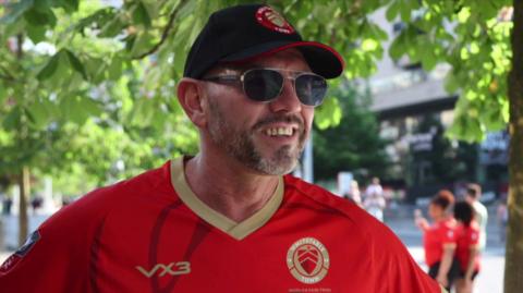 A man with a beard and sunglasses wearing a black Whitstable Town FC hat, sunglasses and a red football shirt.
