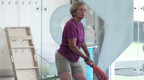A woman holds a red padel bat as she waits to be served a ball.