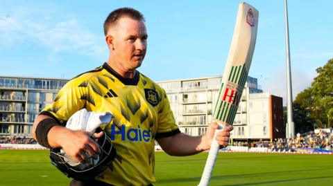 Gloucestershire's D'Arcy Short walks off the pitch at the The Seat Unique Stadium, Bristol, with his bat raised in his left hand and his helmet and gloves under his right arm.
