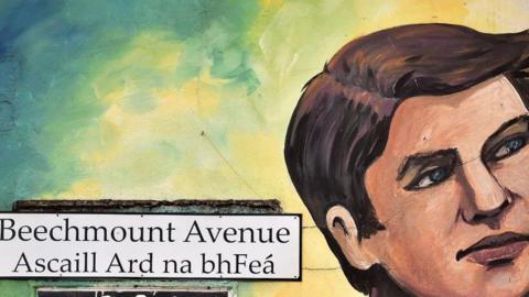 A wall with a white street sign on it saying 'Beechmount Avenue/Ascaill Ard na bhFeá' in black font. The wall is displaying a mural of a face looking to the right.