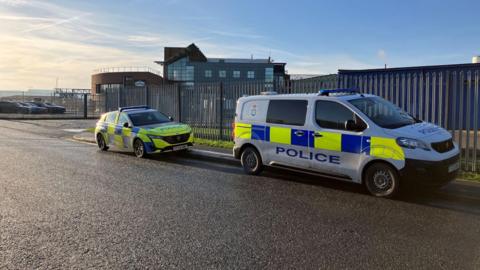 Two marked police vehicles—a car and a van with yellow and blue checkered livery—are parked beside a tall metal fence near a factory.