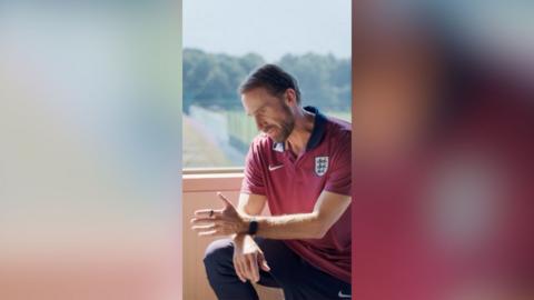 A man in burgundy football shirt sits by a window and looks off to one side