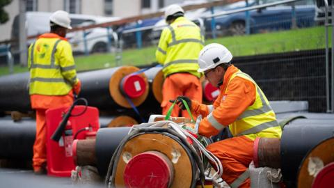 Three men wearing high viz clothing and white construction helmets are working on large pipes, perhaps a metre in diameter, which appear to be largely made of foam insulating material. 