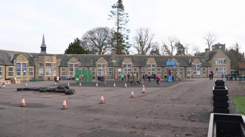 Sele First school, which is a long, single storey, older building with lots of windows. There are children playing outside in the yard that is dotted with traffic cones and some play equipment made with tyres.