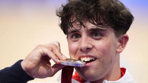 Great Britain's Archie Atkinson celebrates with the silver medal following the 4000m Individual Pursuit gold Final at the National Velodrome on day three of the Paris 2024 Summer Paralympics. He is biting the medal in celebration. 