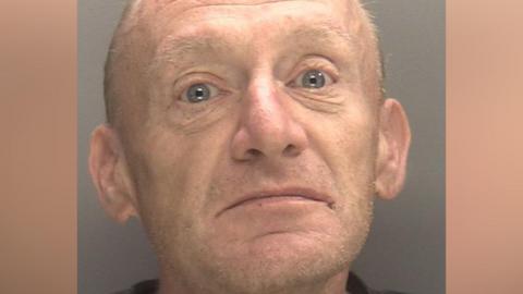 A mug shot of a man with very short light brown hair and facial stubble. He is wearing a dark coloured top