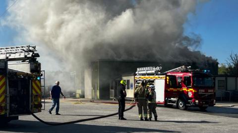 Two fire engines can be seen, with one on the left and another to the right, with a plume of smoke emerging from a building in the background while in the foreground three firefighters can be seen, with a man in blue jeans and a shirt walking toward them from the left. 