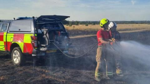 Two firefighters wearing helmets stand in front of an estate vehicle. a water hose is attached to it and they are spraying water onto charred grass at a fire in Rainham in July 2025.