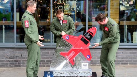 Three RAF pilots in green flying suits stand around an ice carving of a Red Arrows jet located outside a takeaway restaurant