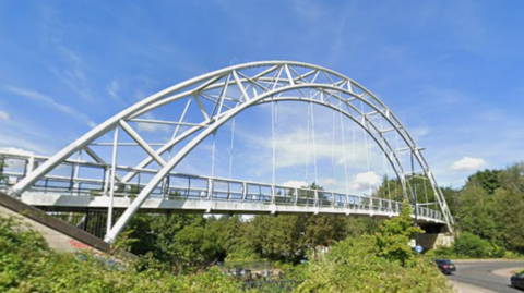 A white metal pedestrian bridge, which has a large arch over it, is suspended over a road in a residential area, surrounded by trees. There are cars coming off a roundabout to drive underneath it. The sky above is bright blue with a few wispy white clouds.
