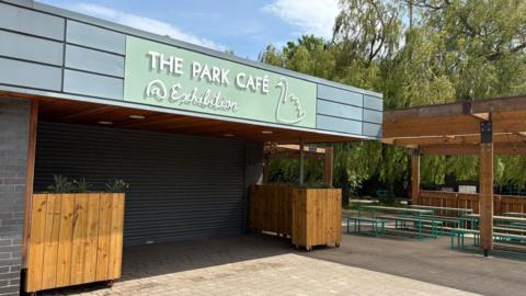 The exterior of The Park Cafe in Exhibition Park, which had a pastel green sign with the outline of a swan on it. The shutters are closed over the entrance. There are planters either side of the entrance and benches underneath an outdoor canopy made from wood.