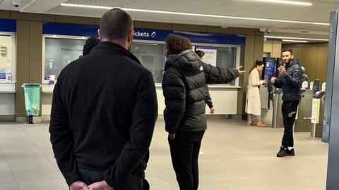 A man in a black coat with curly hair is making a hand gesture clearly annoyed to a group of people.