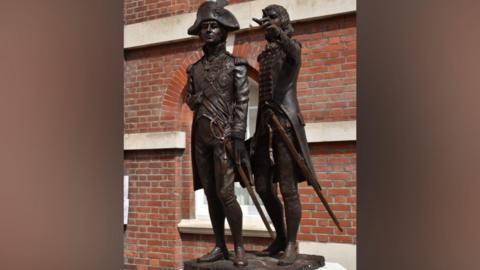 A statue features two figures both in uniform. The statue stands in front of a red brick building with an arched window