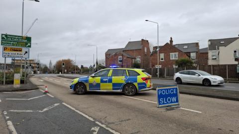 A police road block with a car parked across the approach to the bridge and a blue Police Slow sign set up