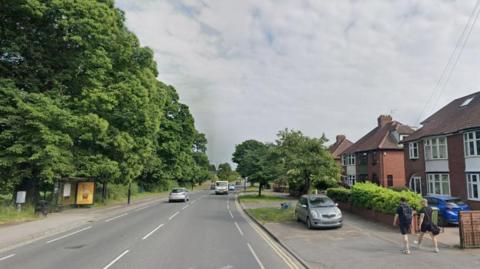 A road with large trees on one side and houses on the other, with a few cars and two men walking on the pavement