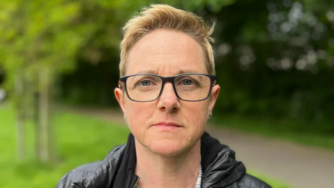 Roz Adams, wearing glasses and looking straight into the camera. She is standing in a park.