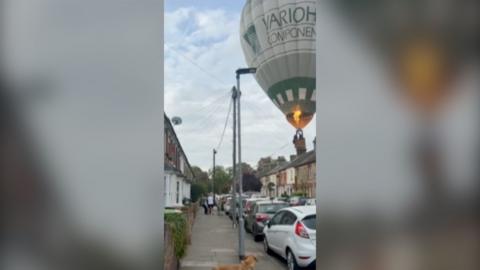 Hot air balloon in the sky seen approaching residential street. Houses on both sides and cars parked in front.