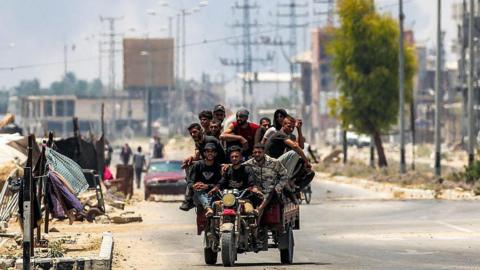 Men ride on a motorised cart along Salah al-Din road in Deir al-Balah, as smoke billows in the background, in the central Gaza Strip (21 July 2025)